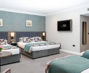 Spacious hotel room with three beds, elegant decor, wall art, flat-screen TV, accent lighting, comfy seating, warm ambience.