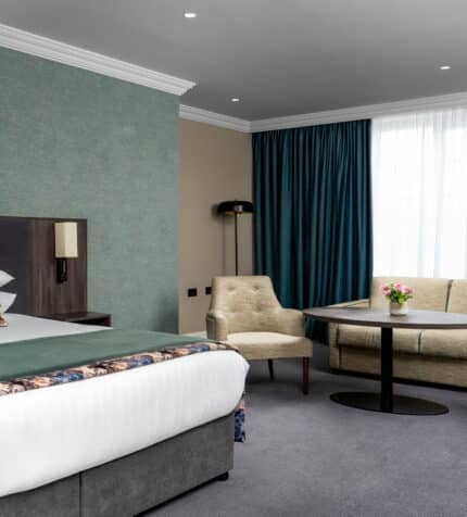 Spacious hotel room with king-size bed, elegant decor, cosy seating area, and large windows offering natural light.