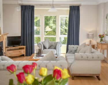 Elegant hotel lounge with plush grey sofas, large windows with garden view, and fresh roses on a wooden table. Relax in style.
