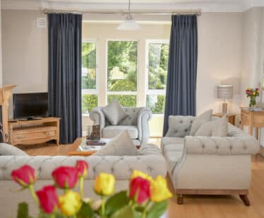 Elegant hotel lounge with plush grey sofas, large windows with garden view, and fresh roses on a wooden table. Relax in style.
