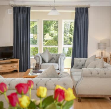 Elegant hotel lounge with plush grey sofas, large windows with garden view, and fresh roses on a wooden table. Relax in style.