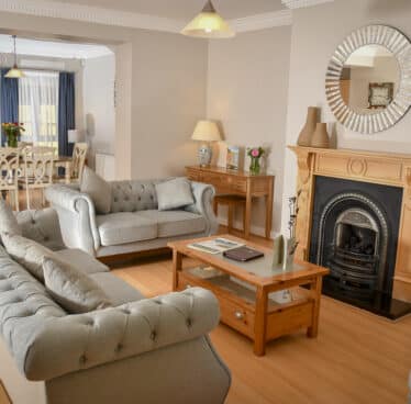 Elegant living room with plush sofas, a cosy fireplace, wooden floors, natural light, and a dining area with flowers.