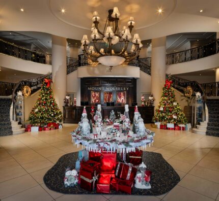 Elegant hotel lobby with festive decorations, grand chandelier, twin staircases, and Christmas trees with presents.
