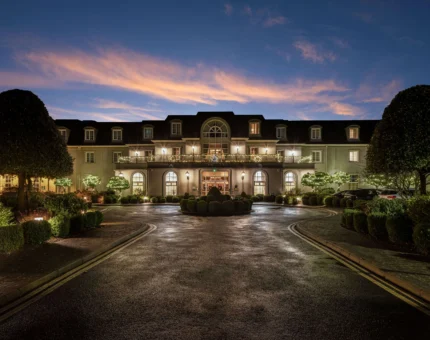 Grand hotel entrance at dusk, elegant architecture, lush gardens, warm lighting; luxurious and inviting stay.
