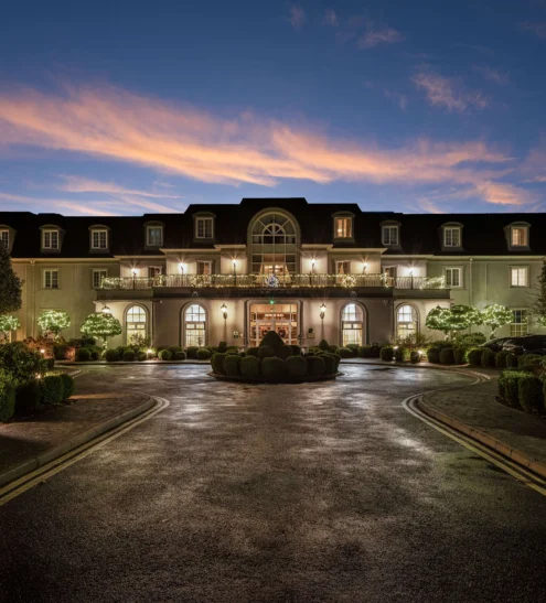Grand hotel entrance at dusk, elegant architecture, lush gardens, warm lighting; luxurious and inviting stay.