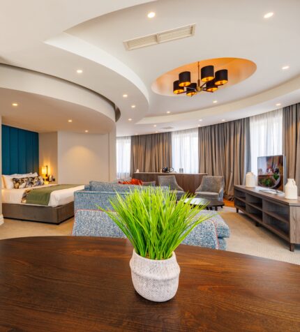 Spacious hotel suite with comfortable bed, modern decor, plush sofas, flat-screen TV, and natural light from large windows.