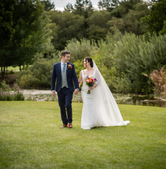 Newlyweds walk hand in hand on lush green lawn by serene pond, surrounded by vibrant trees at a picturesque countryside hotel.