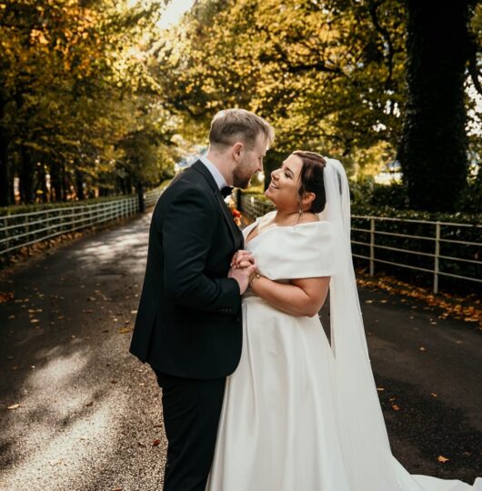 Happy couple on forested driveway, bride in elegant gown, groom in sharp suit, lush foliage surrounds, perfect for weddings.