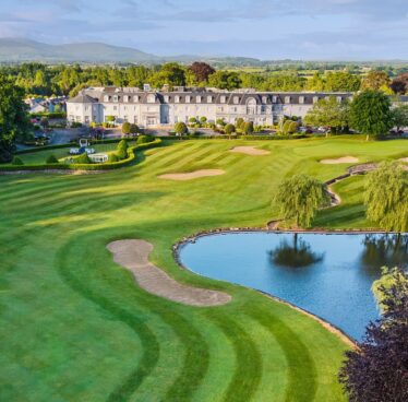 Elegant hotel with adjacent lush golf course, serene pond, surrounded by vibrant greenery and scenic countryside views.