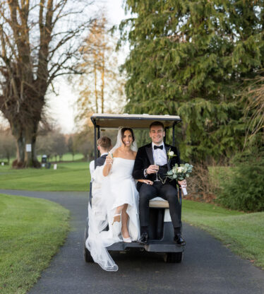 Smiling couple in wedding attire on a golf buggy, surrounded by lush greenery at a picturesque hotel estate.