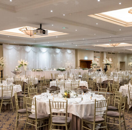Elegant hotel ballroom set for a wedding with round tables, floral centrepieces, and warm lighting.