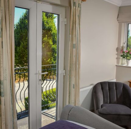 Elegant hotel room with French doors opening to a garden view, featuring a plush grey chair and a classic wooden desk.