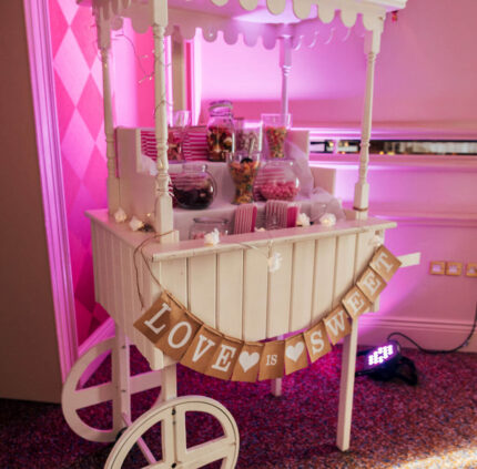 Charming white sweet cart with jars of colourful treats, set in a cosy, pink-themed hotel corner for a delightful stay.
