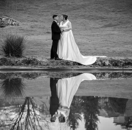 A newlywed couple embraces by a serene pond, lush green grounds surrounding them, reflecting romance and tranquility.