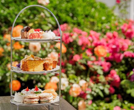 Afternoon tea stand with cakes and sandwiches on a garden table, vibrant flowers in the background.