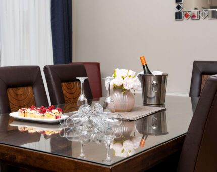 Elegant dining area with glass table, plush chairs, fresh flowers, wine, and gourmet cakes, creating a luxurious experience.