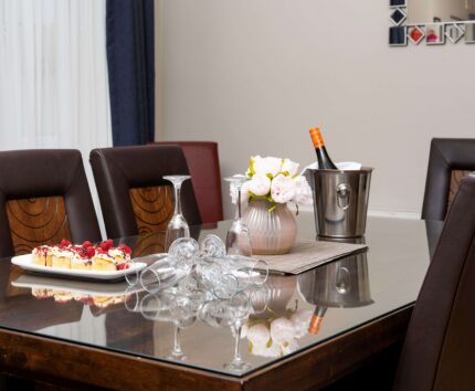 Elegant dining area with glass table, plush chairs, fresh flowers, wine, and gourmet cakes, creating a luxurious experience.