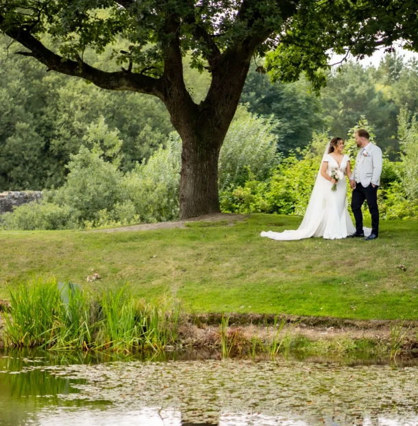 Couple on riverside lawn beneath a large tree, perfect for wedding ceremonies, surrounded by greenery and pond reflections.