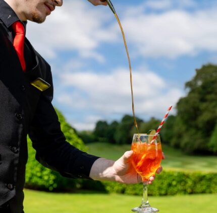 A waiter elegantly pours a refreshing cocktail in a sunny garden, surrounded by lush greenery at a luxury hotel.