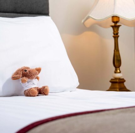 Cozy hotel room with plush bedding, elegant lamp, and charming toy on the bed showcasing comfort and warmth.