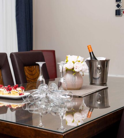Elegant hotel dining area with a glass table set for wine and dessert, adorned with flowers, offering a luxurious stay.